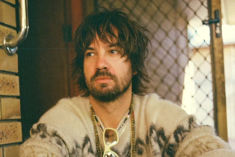 Kevin Parker of Tame Impala wearing a patterned sweater, beaded necklaces, and sunglasses indoors in a casual portrait.